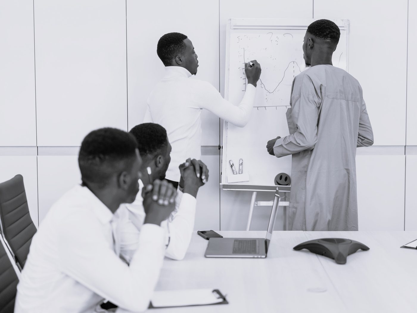 Four colleagues around a boardroom table, one sketching a chart on a flip chart while the others follow