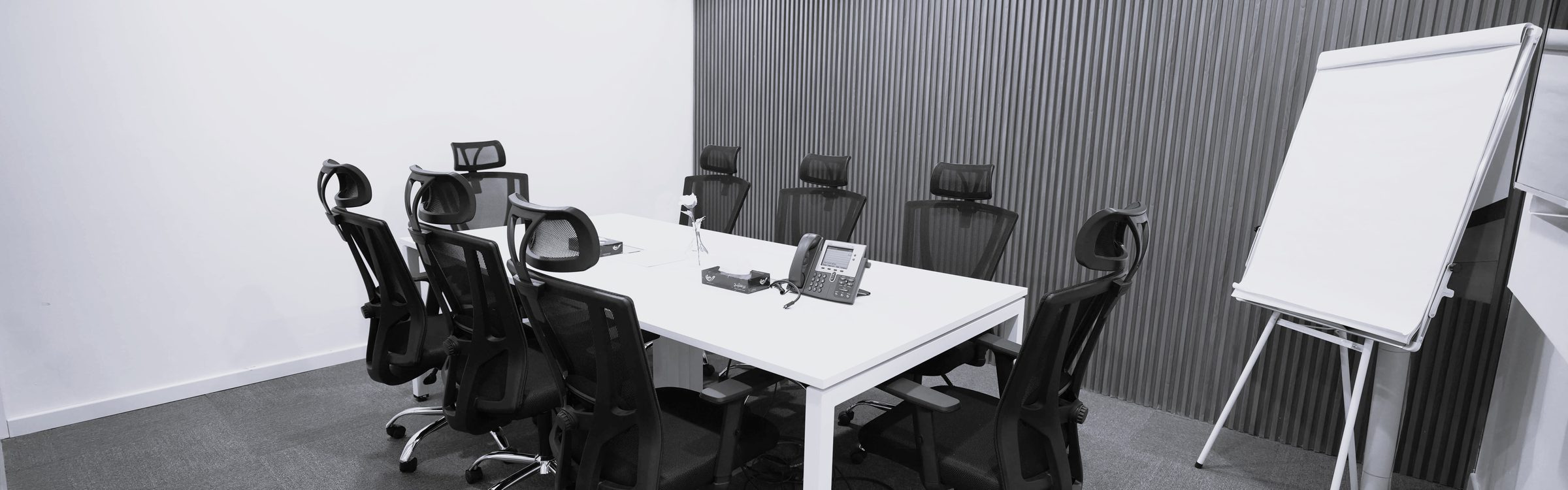 A meeting room with a long white table, black mesh chairs and a flip chart against a slatted wall